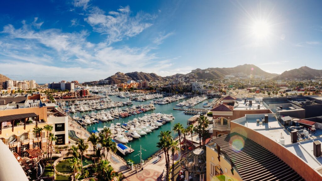 The marina in Cabo, Mexico on a sunny day. Boats lined up along the slip and mountains in the background.